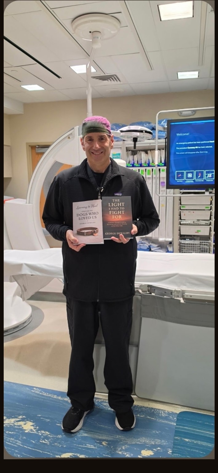 George Dewiliby smiling and holding his two published books in a cardiac catheterization laboratory